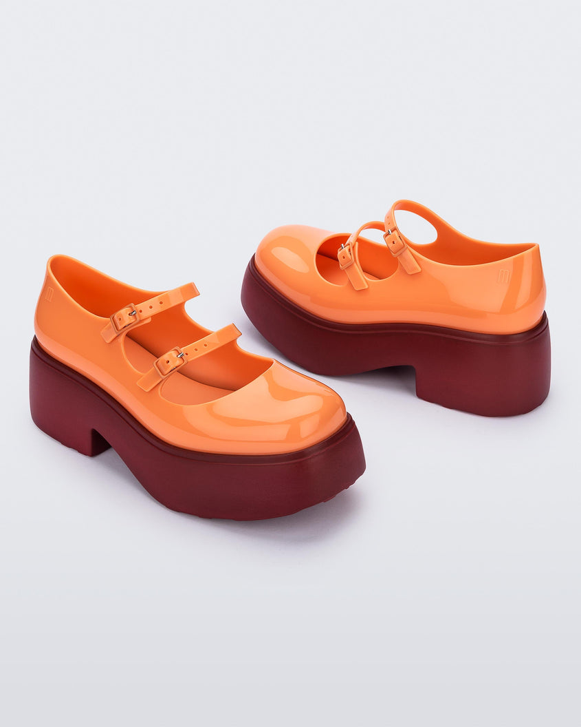 A top and side view of a pair of orange Melissa Farah platform shoes, with a brown sole and two orange straps fastened by orange buckles.
