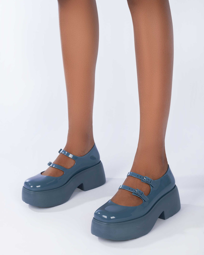 A model's legs wearing a pair of blue Melissa Farah platform shoes with two straps fastened by blue buckles.