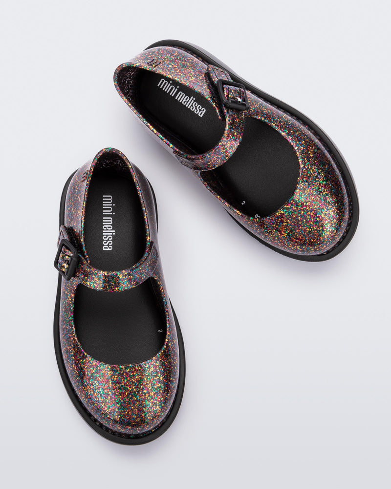 Top view of a pair of clear multicolor glitter Mini Melissa Lola loafer with a rounded toe, strap and black buckle.