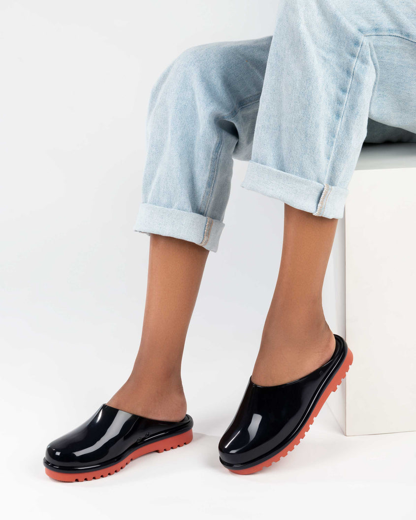 A model's legs in denim, wearing a pair of black Melissa Smart Clogs with a black base and an orange/brown sole.