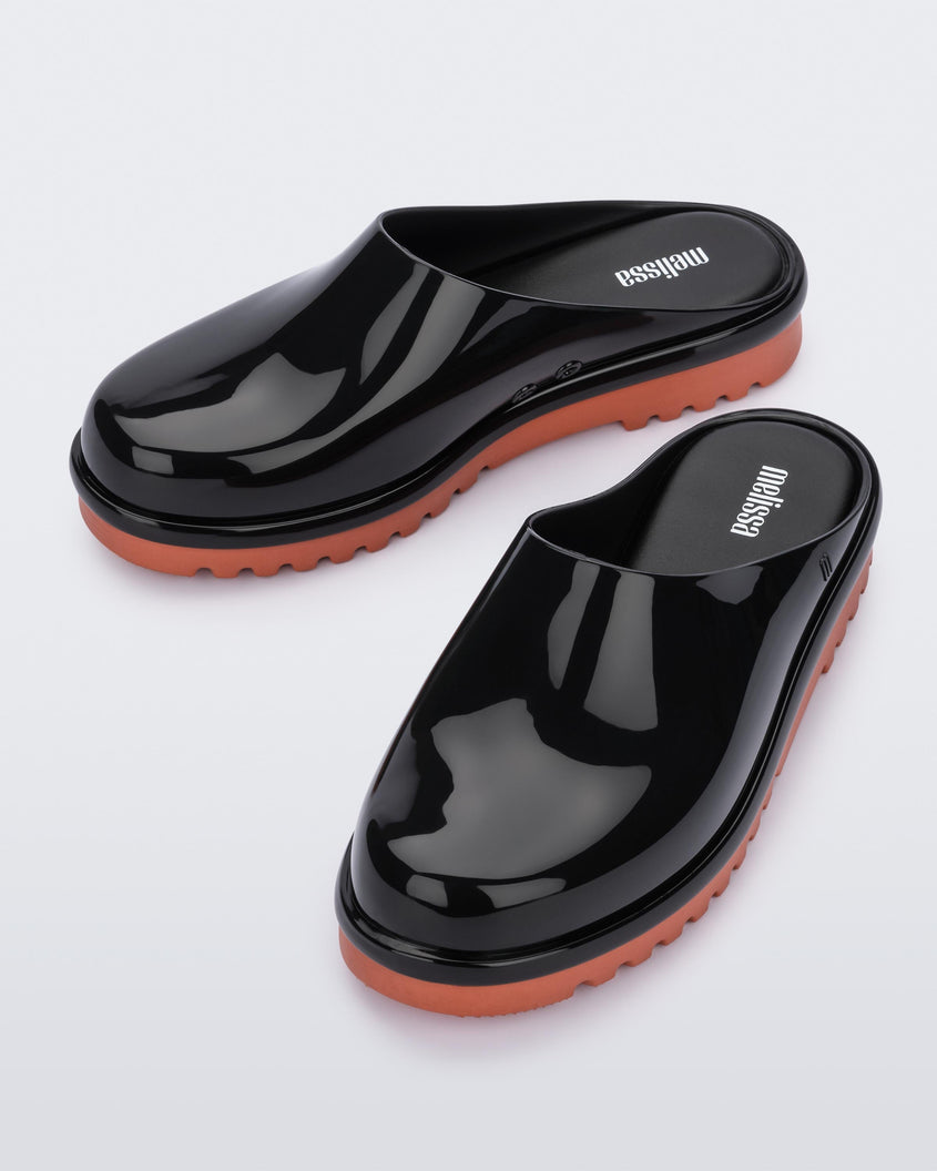 An angled top view of a pair of black Melissa Smart Clogs with a black base and an orange/brown sole.