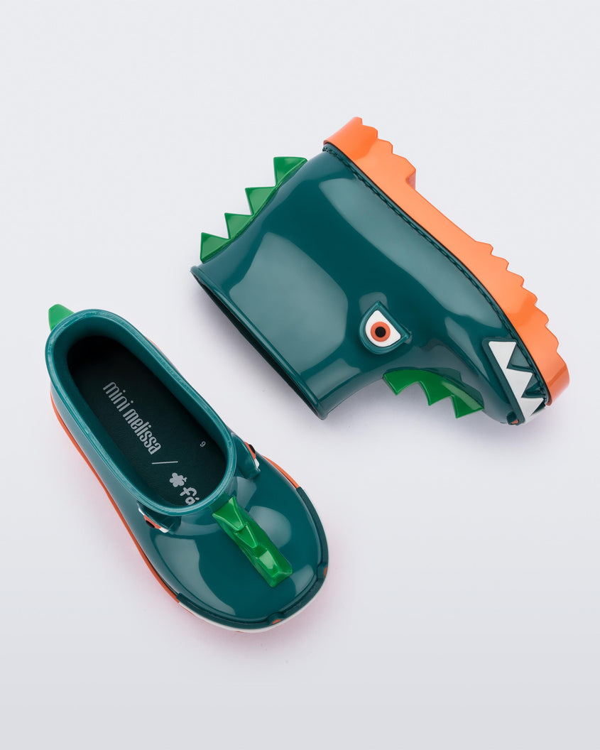 A top and side view of a pair of orange/green Mini Melissa Rain Boots with a green base, orange sole, light green triangle details, white triangle details in the front and a set of orange eyes which looks like a dinosaurs face,