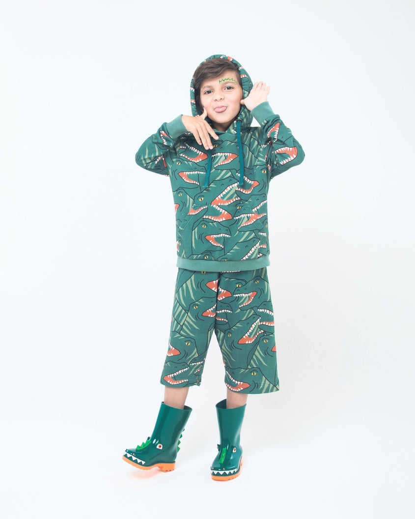 A model posing for a picture, in a green patterned jacket and shorts and a pair of orange/green Mini Melissa Rain Boots with a green base, orange sole, light green triangle details, white triangle details in the front and a set of orange eyes which looks like a dinosaurs face, laying on their side.