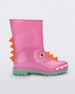 Side view of a green/pink Mini Melissa Rain Boot with a pink base, green sole, orange triangle details, white triangle details in the front and a set of orange eyes which looks like a dinosaurs face,