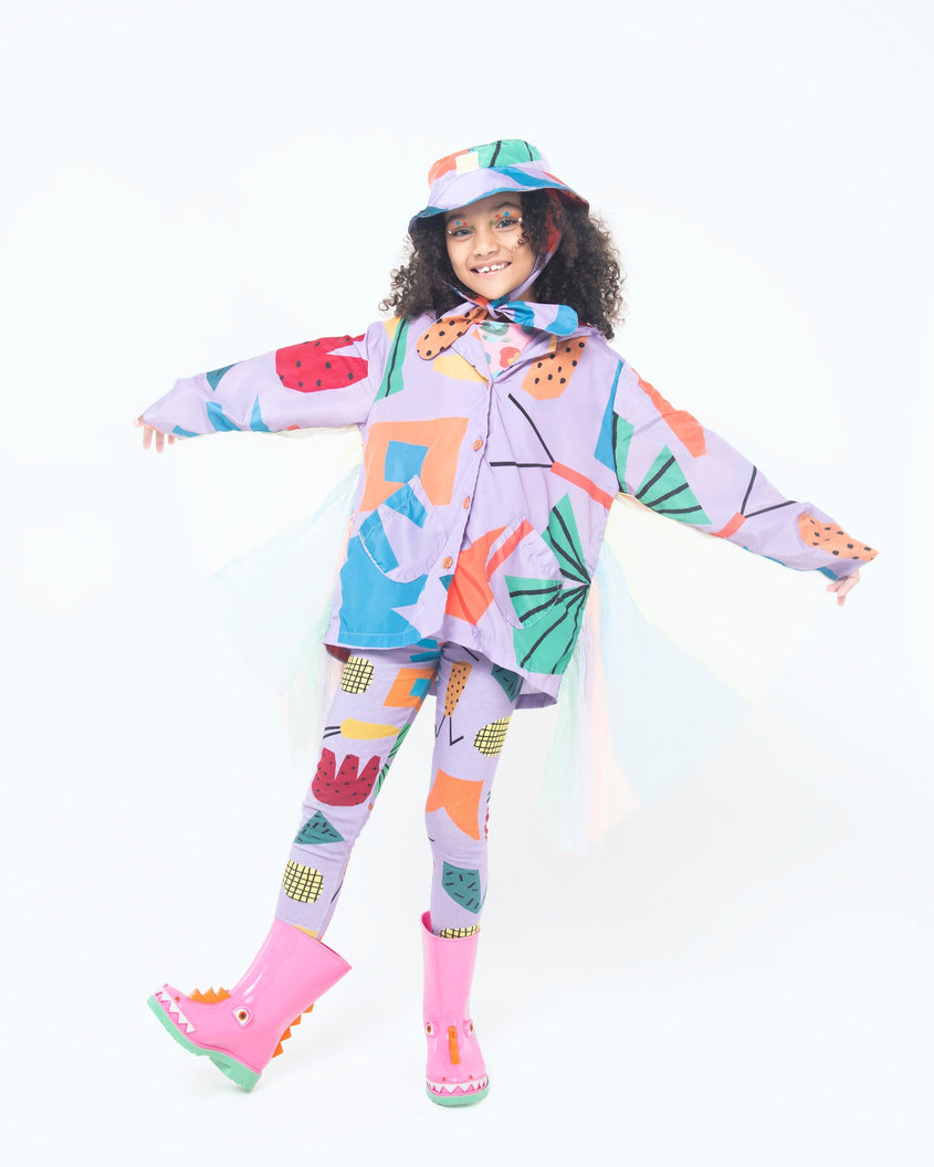 A model posing for a picture wearing in a purple patterned jacket and pants, wearing a pair of green/pink Mini Melissa Rain Boot with a pink base, green sole, orange triangle details, white triangle details in the front and a set of orange eyes which looks like a dinosaurs face,