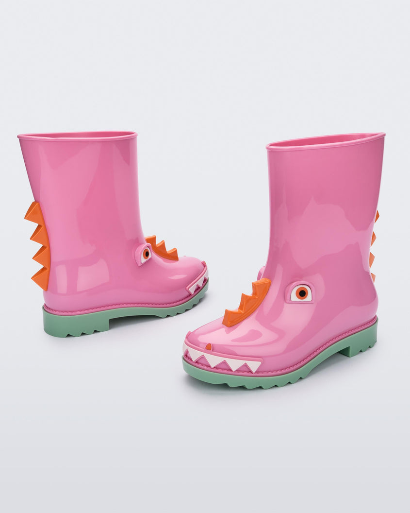 An angled front and side view of a pair of green/pink Mini Melissa Rain Boots with a pink base, green sole, orange triangle details, white triangle details in the front and a set of orange eyes which looks like a dinosaurs face,