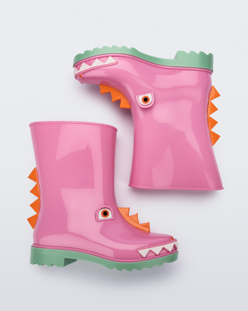 A side view of a pair of green/pink Mini Melissa Rain Boot with a pink base, green sole, orange triangle details, white triangle details in the front and a set of orange eyes which looks like a dinosaurs face, laying on their side.
