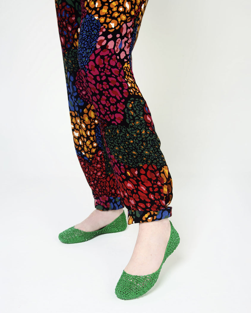 A closeup of a model wearing a pair of Melissa Campana green flats with multi colord printed pants.