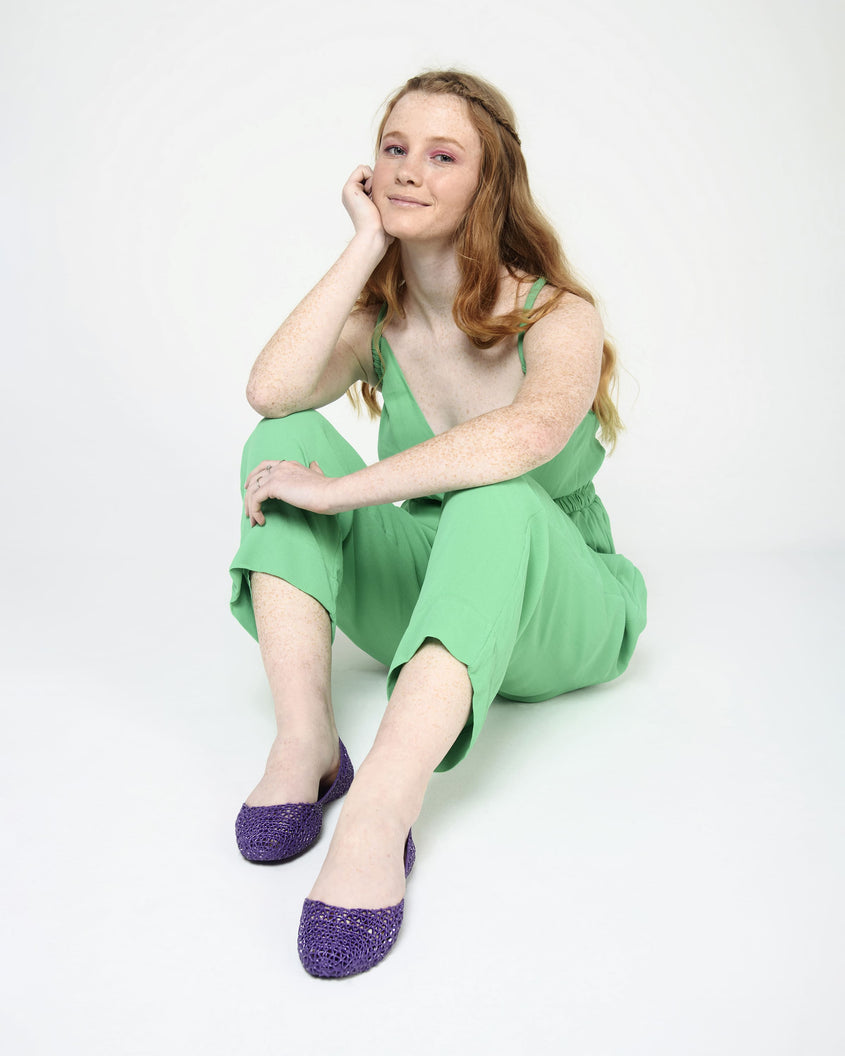 A model sitting and posing in a green jumpsuit wearing a pair of Melissa Campana purple flats with an open woven texture