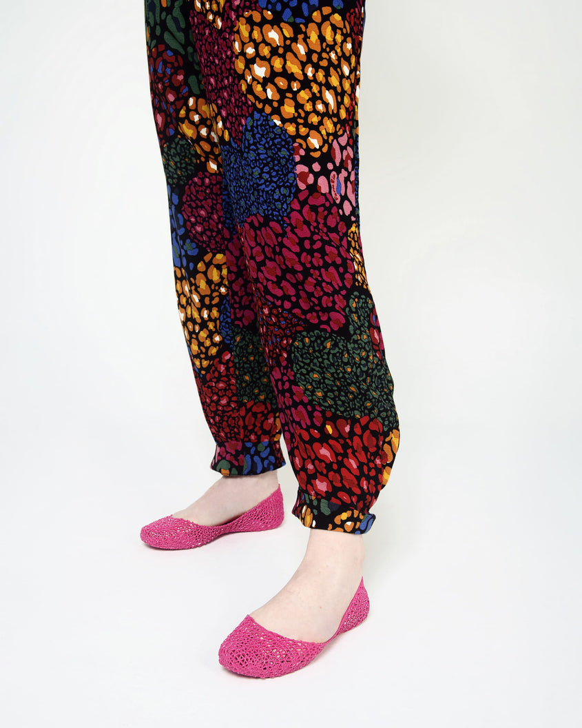 A closeup of a model wearing a pair of Melissa Campana pink flats with multi colord printed pants.