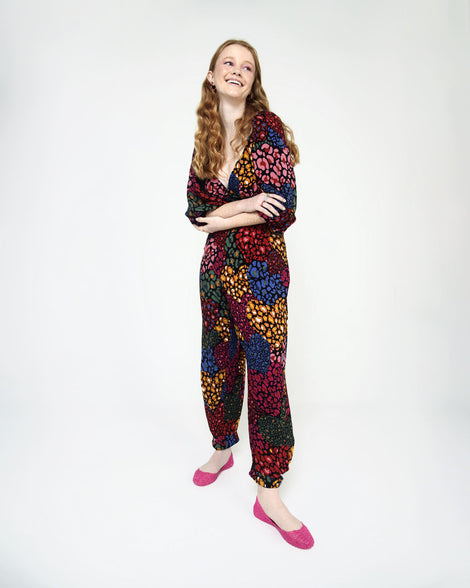 A woman standing with her arms crossed wearing a multicolored pattern jumpsuit with a pair of bright pink Melissa Campana flats. 