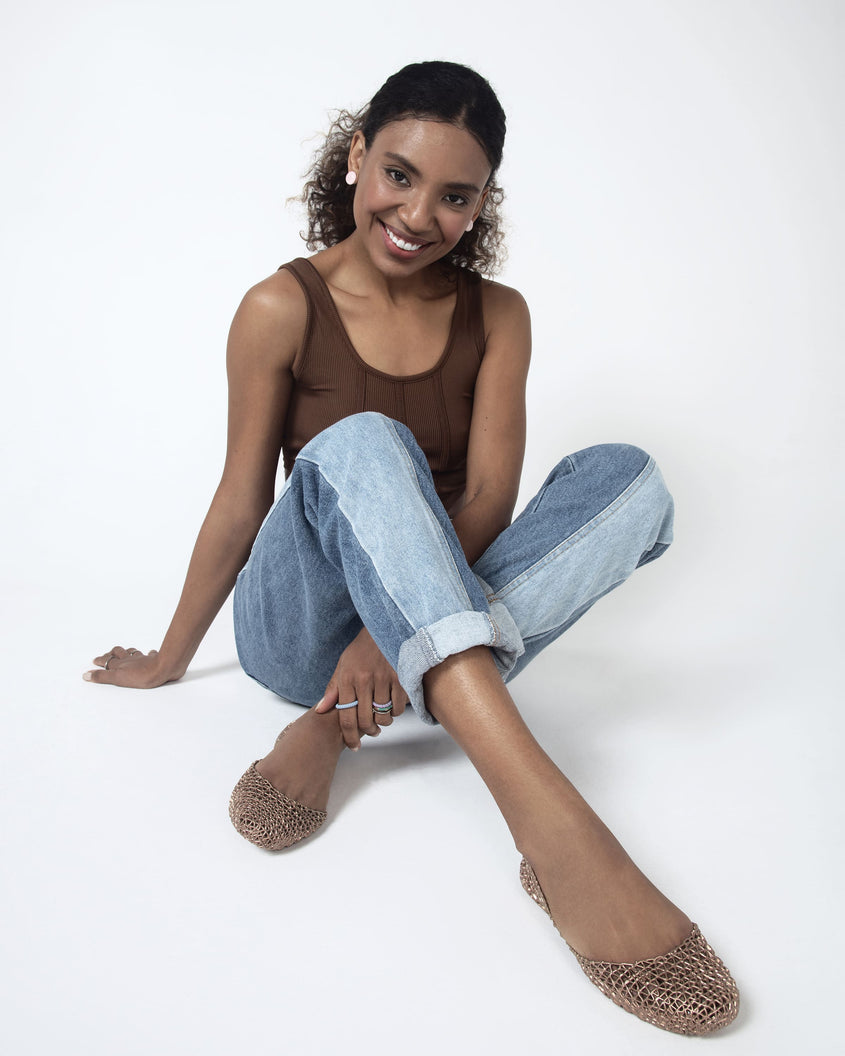 A woman sitting modeling a pair of Melissa Campana flats in gold paired with cuffed jeans and a brown tank top. 