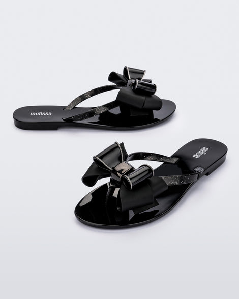 An angled side and front view of a pair of black Melissa Harmonic Sweet flip flops with a black and glitter bow.