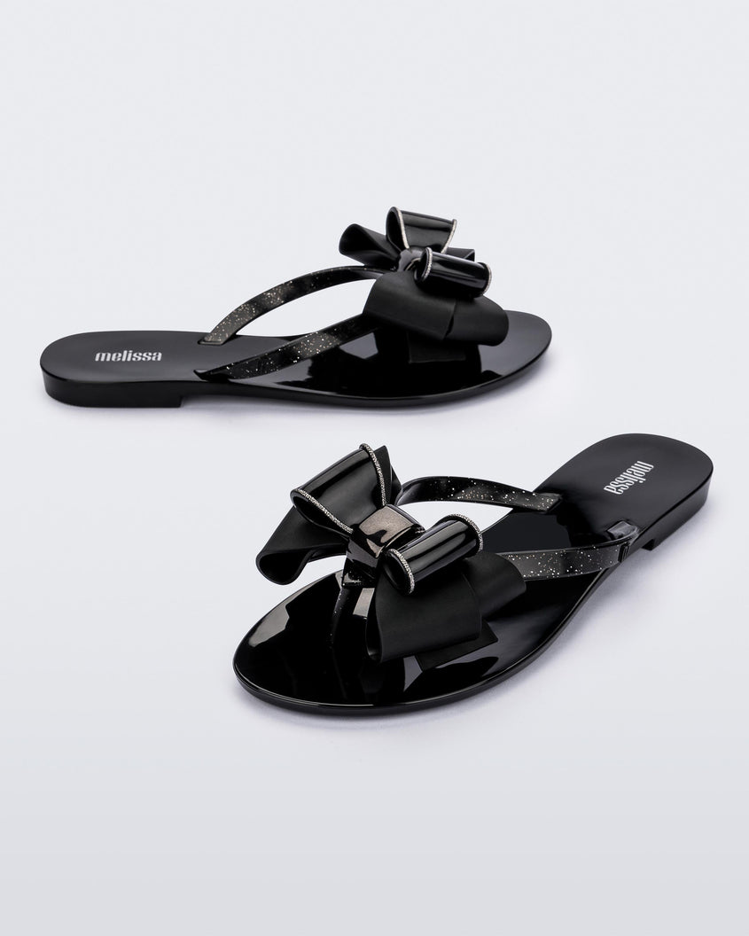 An angled side and front view of a pair of black Melissa Harmonic Sweet flip flops with a black and glitter bow.