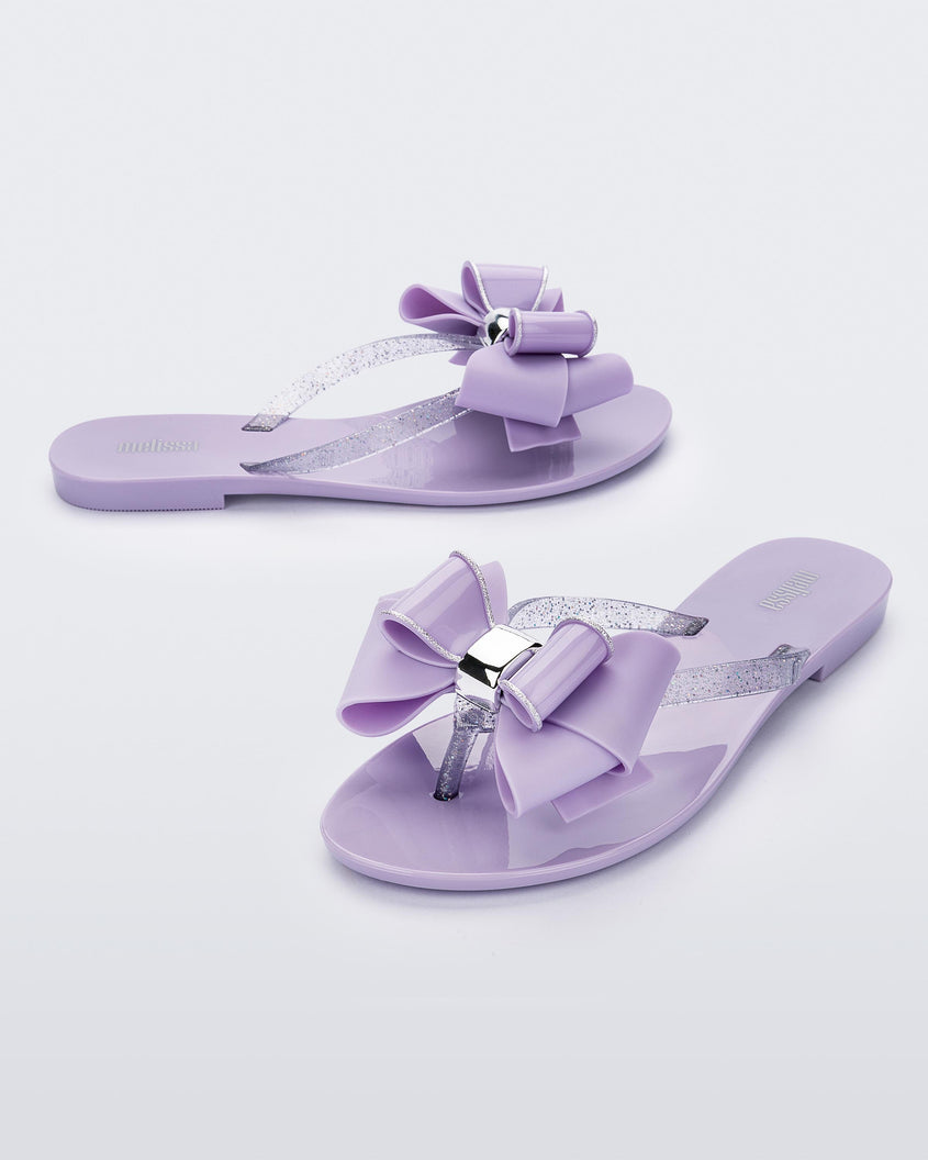 An angled front and side view of a pair of lilac Melissa Harmonic Sweet flip flops with a lilac bow and clear glitter straps.