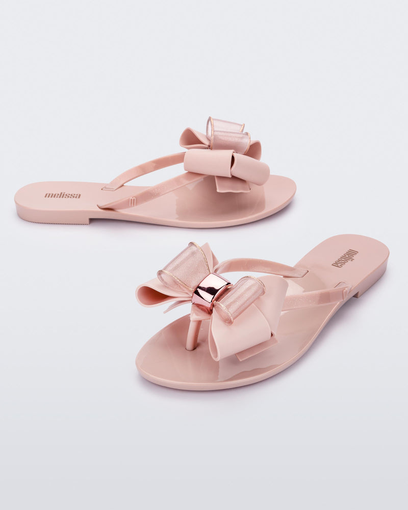 An angled side and front view of a pair of light pink Melissa Harmonic Sweet flip flops with a light pink and glitter bow.