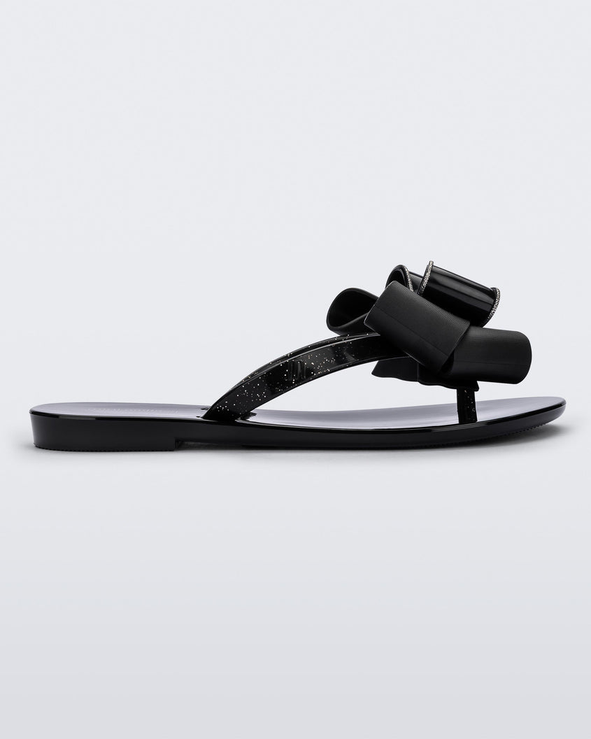 Side view of a black Mini Melissa Harmonic Sweet flip flop with a glitter and black bow detail on the straps.