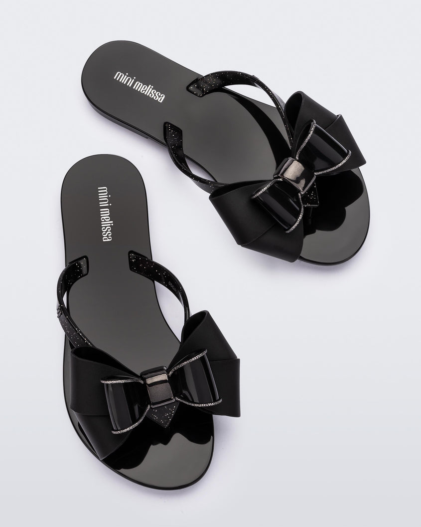 A top view of a pair of black Mini Melissa Harmonic Sweet flip flops with a glitter and black bow detail on the straps.