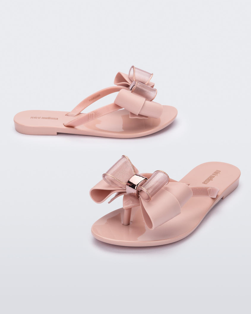 An angled front and side view of a pair of light pink Mini Melissa Harmonic Sweet flip flops with a light pink and glitter bow.
