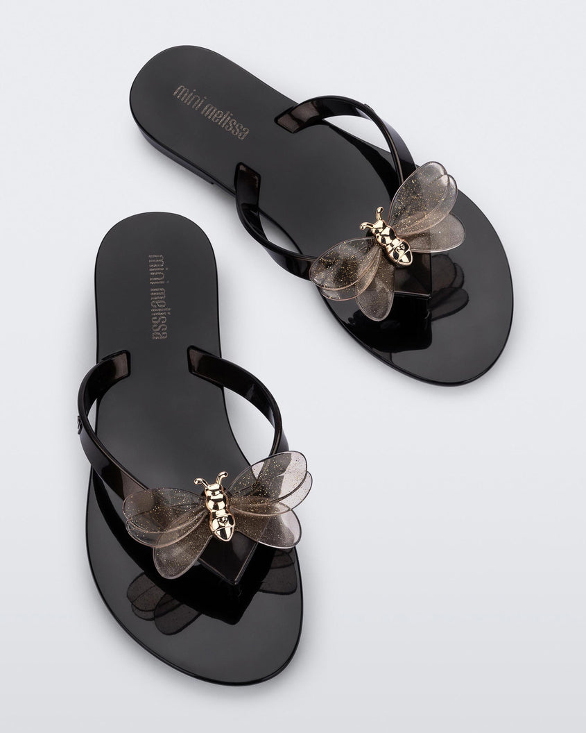 Top view of a pair of black / metallic black Mini Melissa's Harmonic Bugs flip flop with a clear glitter bug with a gold buckle on the straps.