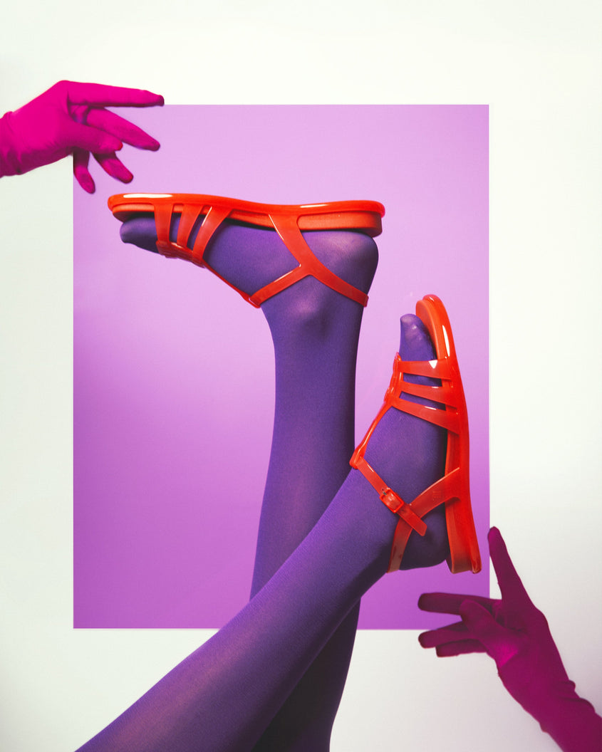 A artistic shot of a models legs in purple tights wearing a pair of red Melissa Party sandals with back ankle strap and buckle closure.
