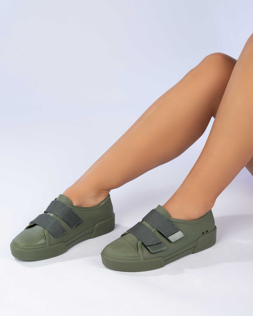 A model's legs wearing a pair of green/orange Melissa Cool sneakers with an orange insole, green base and two green velcro straps.