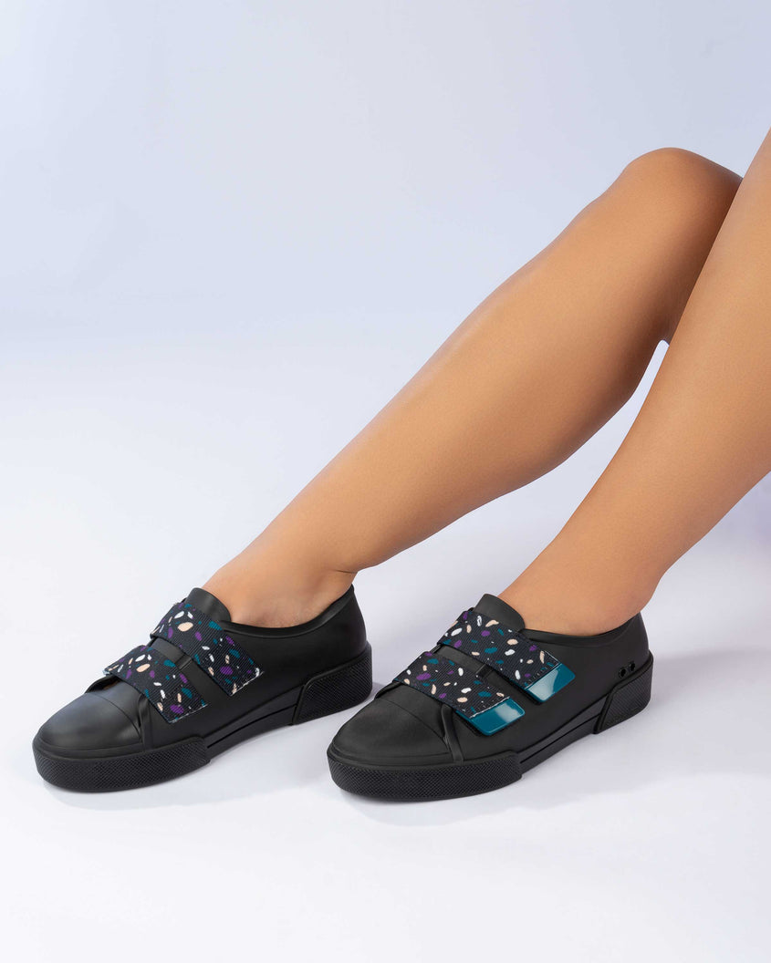 A model's legs wearing a pair of black/purple Melissa Cool sneakers with a purple insole, black base and two black, blue, purple and beige patterned velcro straps.
