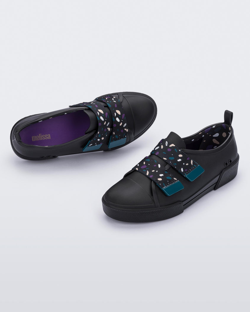 An angled top and side view of a pair of black/purple Melissa Cool sneakers with a purple insole, black base and two black, blue, purple and beige patterned velcro straps.