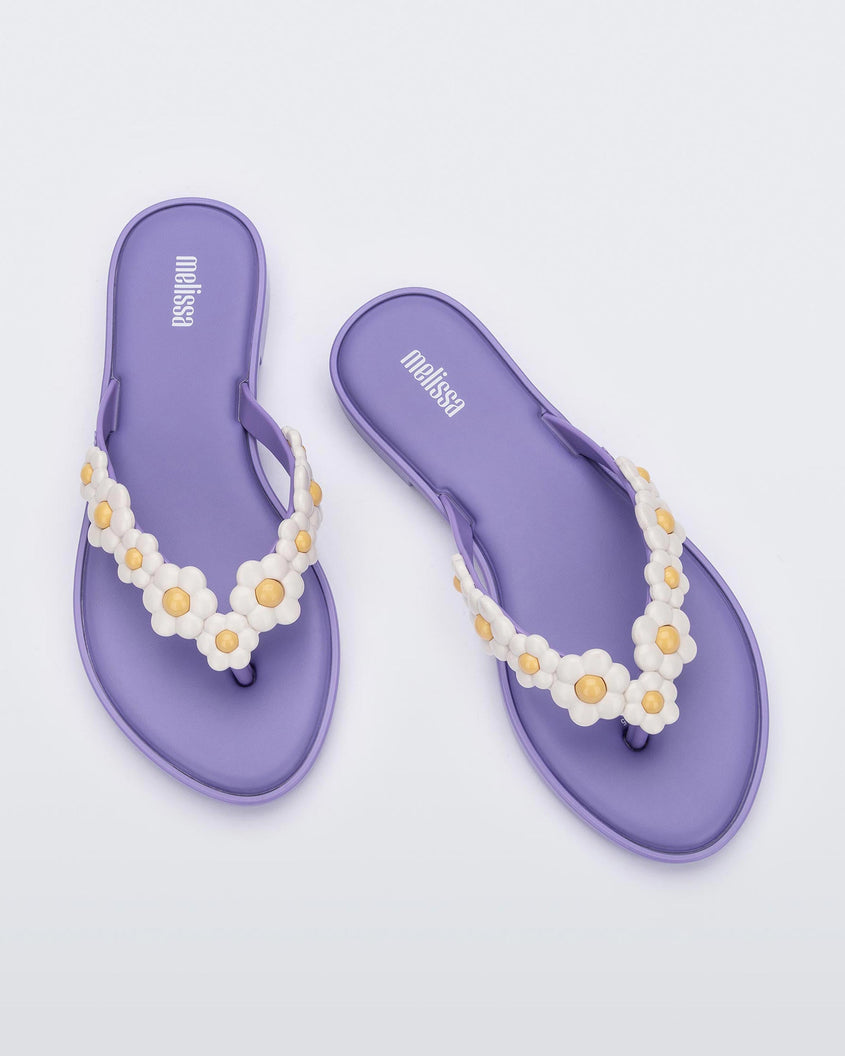 A top view of a pair of lilac / white Melissa Spring Flip Flops with white and yellow flowers on them.