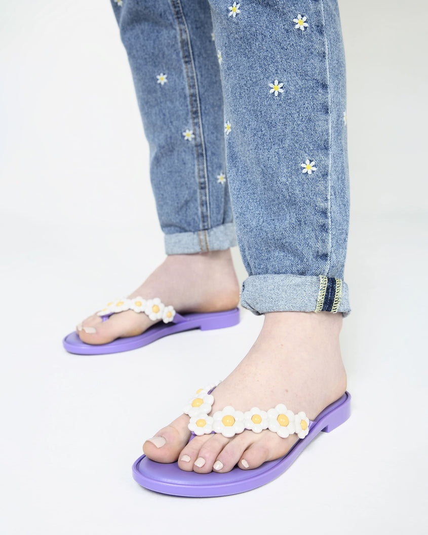 A model's legs wearing a pair of lilac / white Melissa Spring Flip Flops with white and yellow flowers on them.