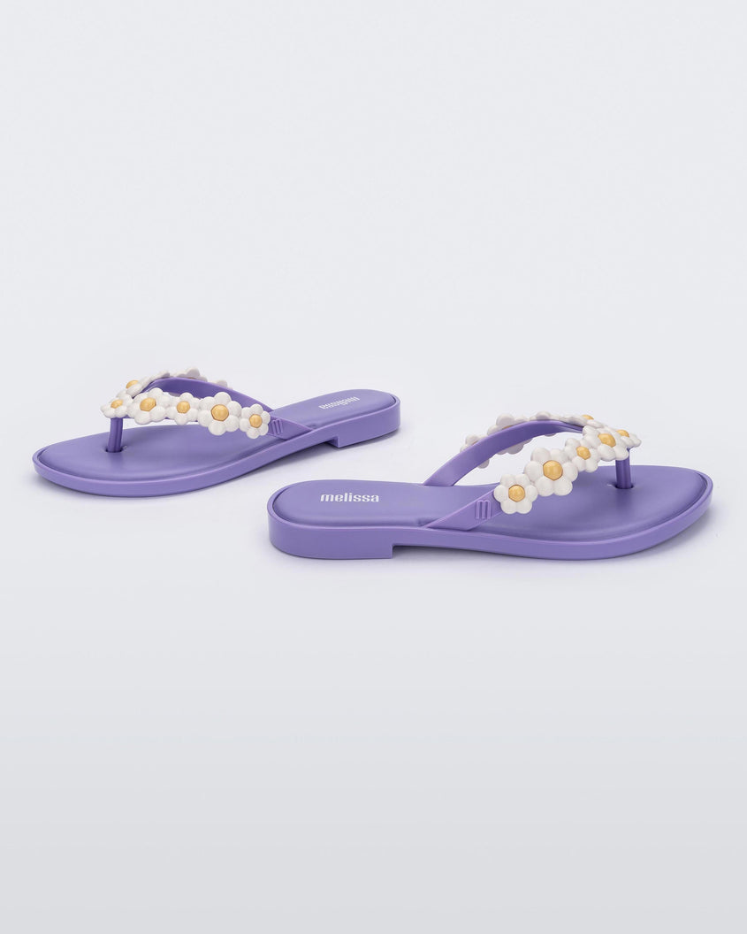 A side and angled view of a pair of lilac / white Melissa Spring Flip Flops with yellow and white flowers on them.