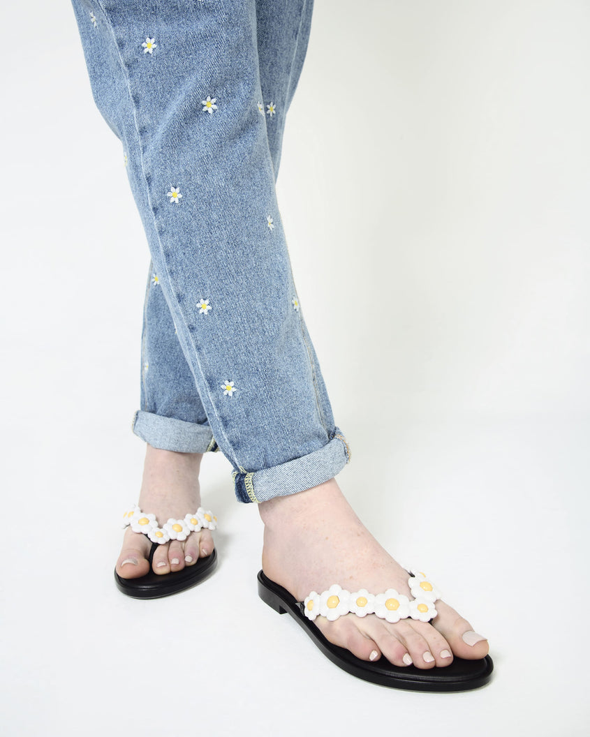 A model's legs wearing a pair of black Melissa Spring Flip Flops with white and yellow flowers on them.