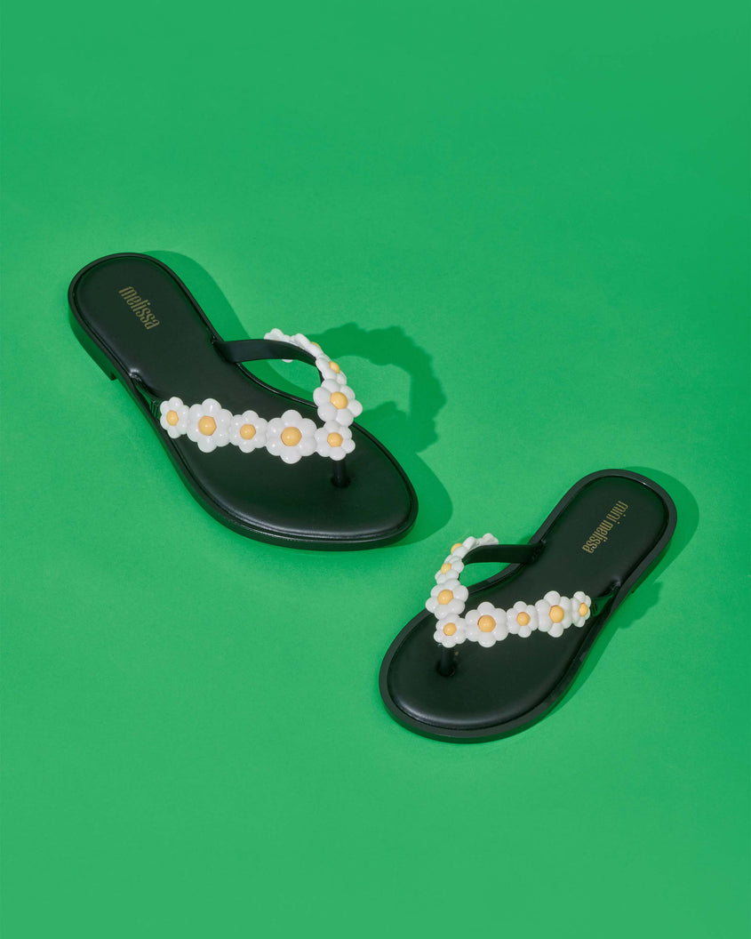 A top and angled view of a pair of black Melissa Spring Flip Flops with white and yellow flowers on them on a green surface.