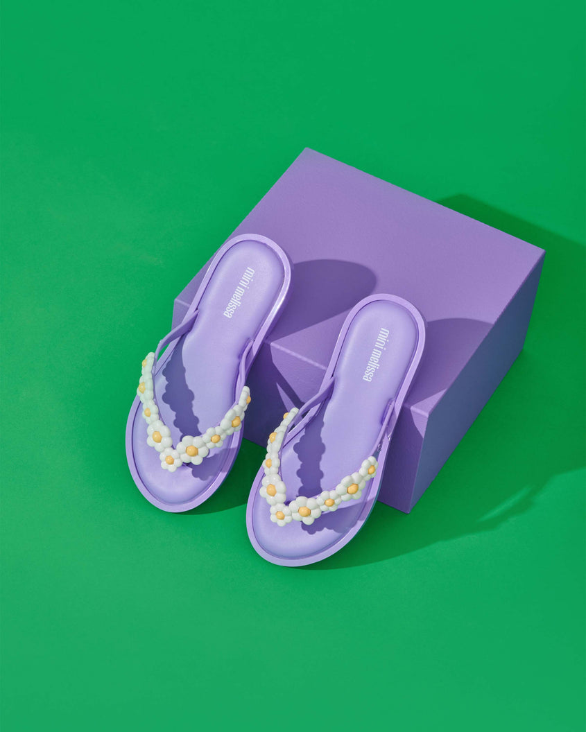 A top view of a pair of lilac Mini Melissa Spring Flip Flops with white and yellow flowers sitting on top of a purple box.