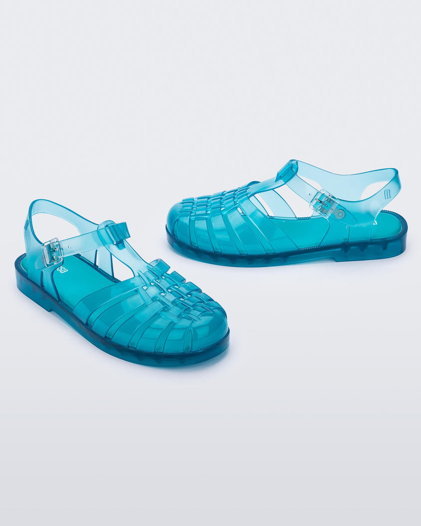 Angled view of a pair of transparent blue Melissa Possession sandals with a closed toe front weft design connected to a top strap with a buckle.