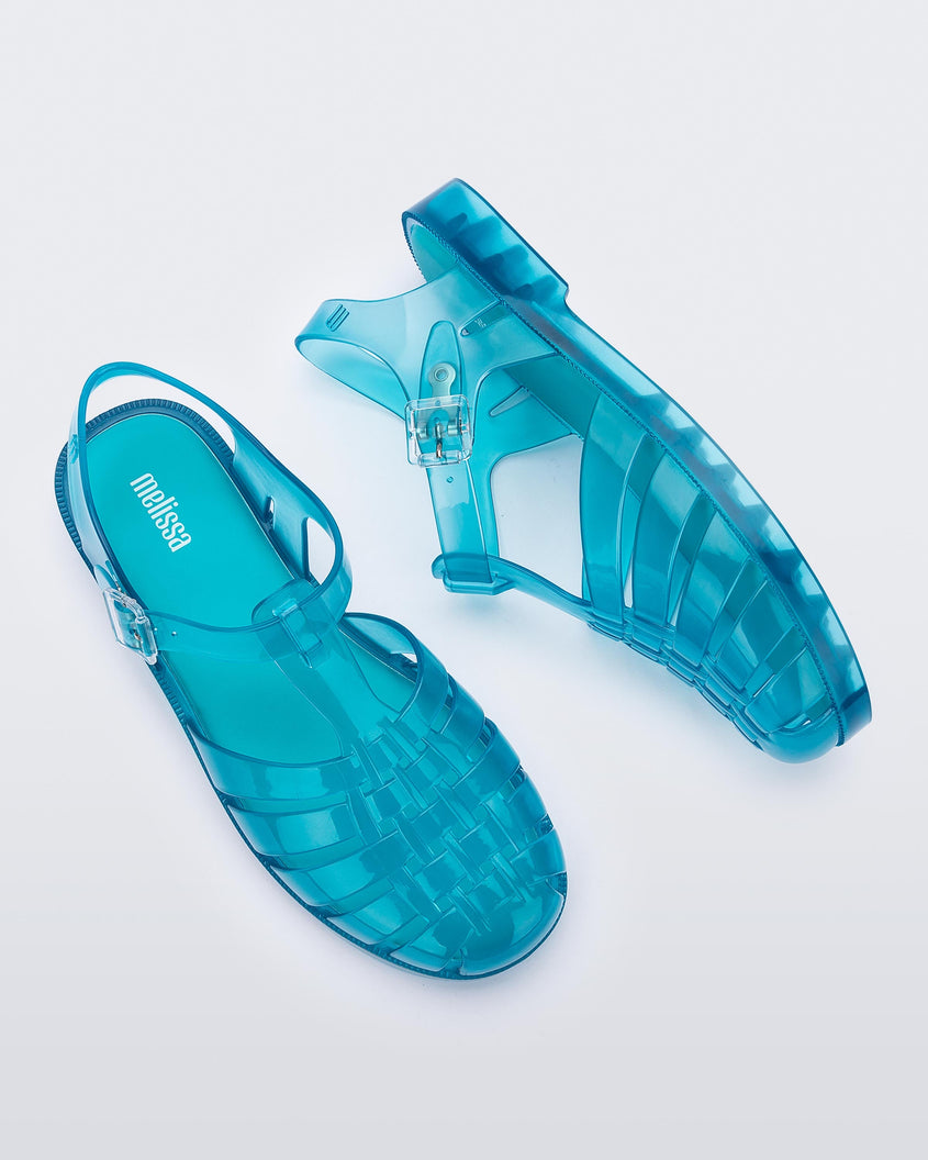 Top and side view of a pair of transparent blue Melissa Possession sandals with a closed toe front weft design connected to a top strap with a buckle.