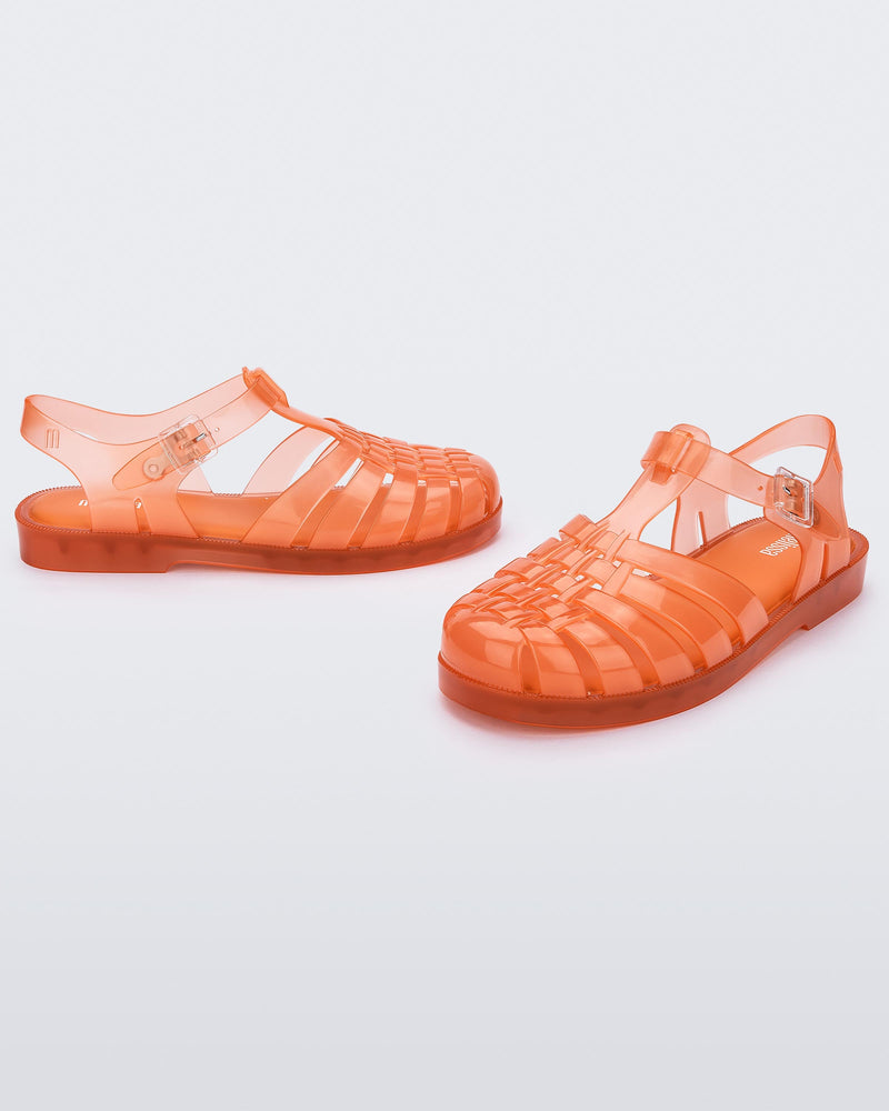 Angled view of a pair of transparent orange Melissa Possession sandals with a closed toe front weft design connected to a top strap with a buckle.