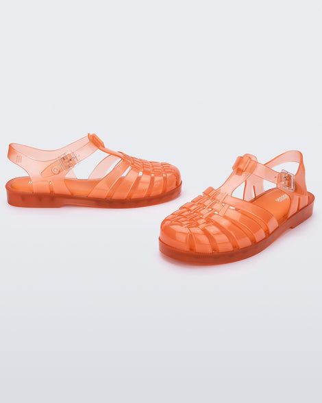 Angled view of a pair of transparent orange Melissa Possession sandals with a closed toe front weft design connected to a top strap with a buckle.