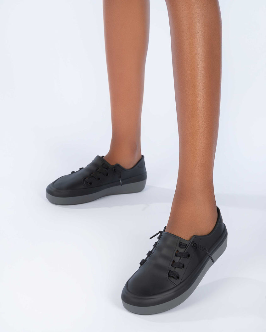 A model's legs wearing a pair of a black Melissa Ulitsa sneakers with a black base, laces and a gray sole.