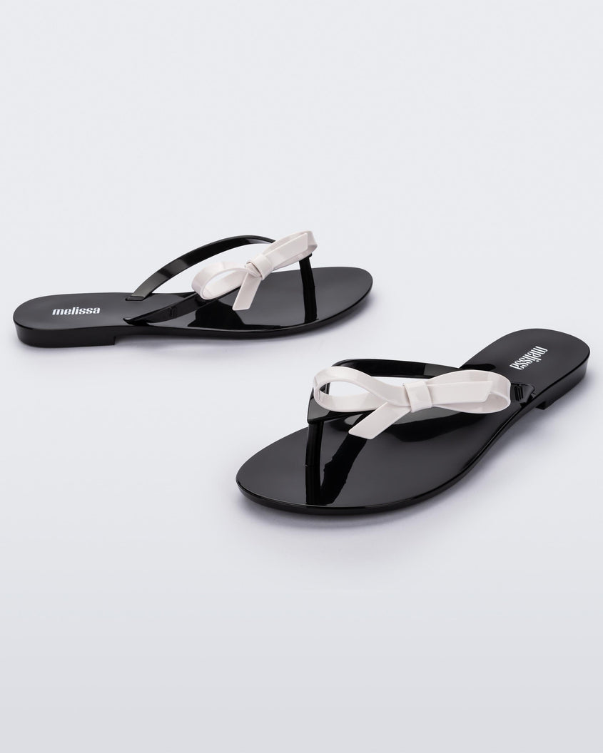An angled front and side view of a pair of black/white Melissa Harmonic Sweet flip flops with a black sole, straps and white bow on top.