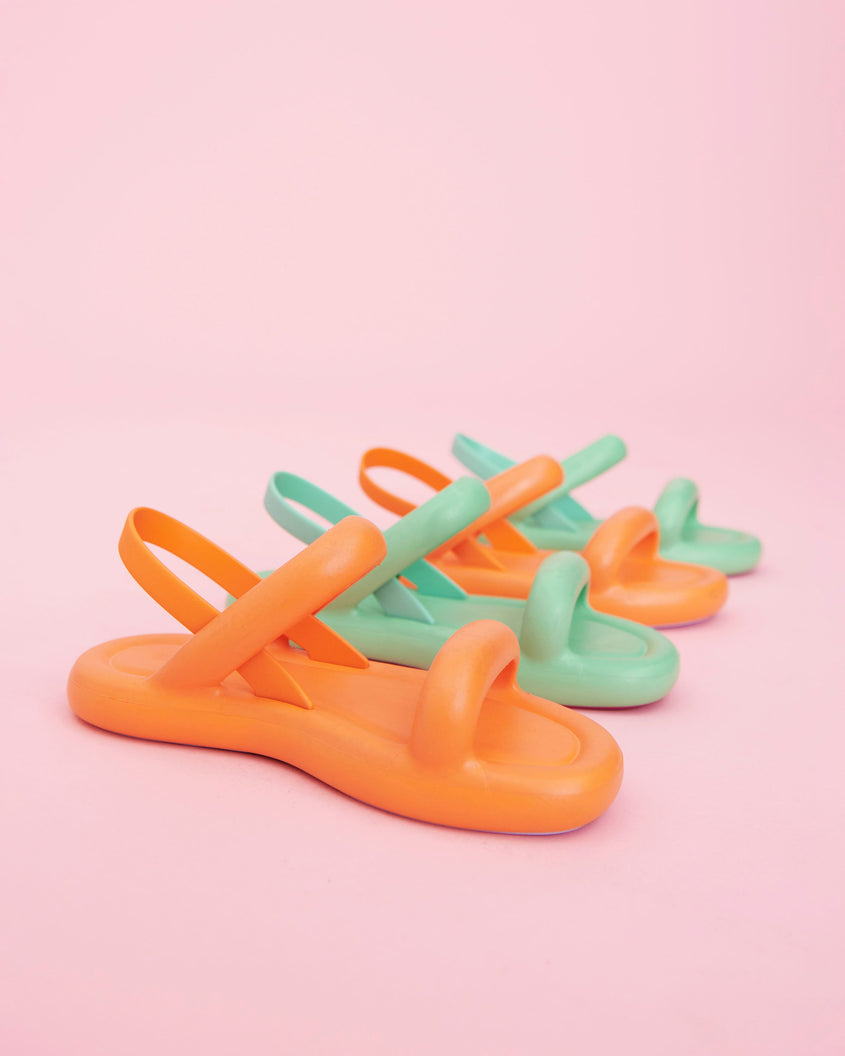 An angled view of a group of Melissa Free Bloom sandals in orange and green, shown alongside eachother on a pink background.