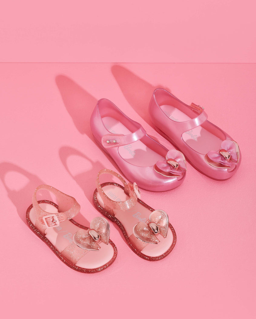 A pair of Mini Melissa Mar Sandals in pink glitter next to a pair of Mini Melissa Ultragirl flats in pink glitter with a bow.
