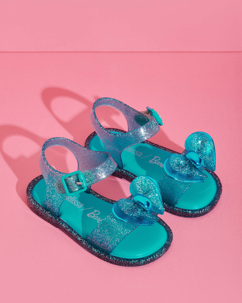 Angled view of a pair of blue glitter Mini Melissa Mar Sandals with bow on a pink background.