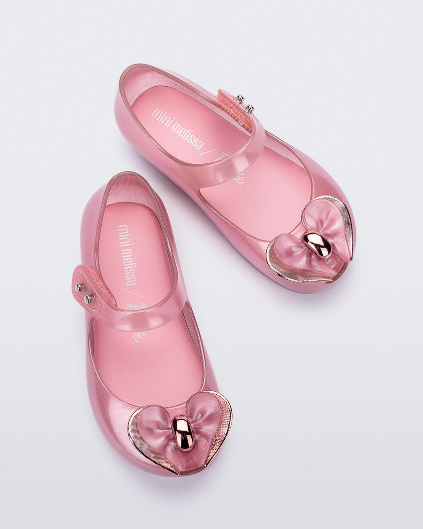Top view of a pair of pearly pink Mini Melissa Ultragirl flats with a top strap and a heart bow buckle detail on the toe.