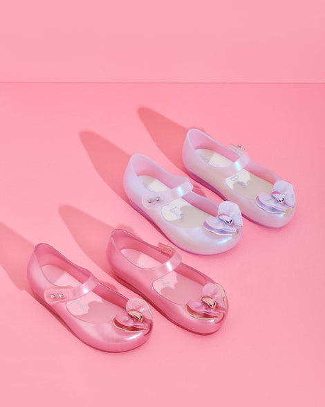 An angled top view of two pairs of pearly pink and pearly blue Mini Melissa Ultragirl flats with a top strap and a heart bow buckle detail on the toe.