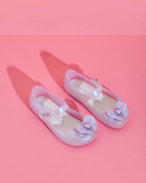 An angled top view of a pair of pearly blue Mini Melissa Ultragirl flats with a top strap and a heart bow buckle on the toe, on top of a pink surface.