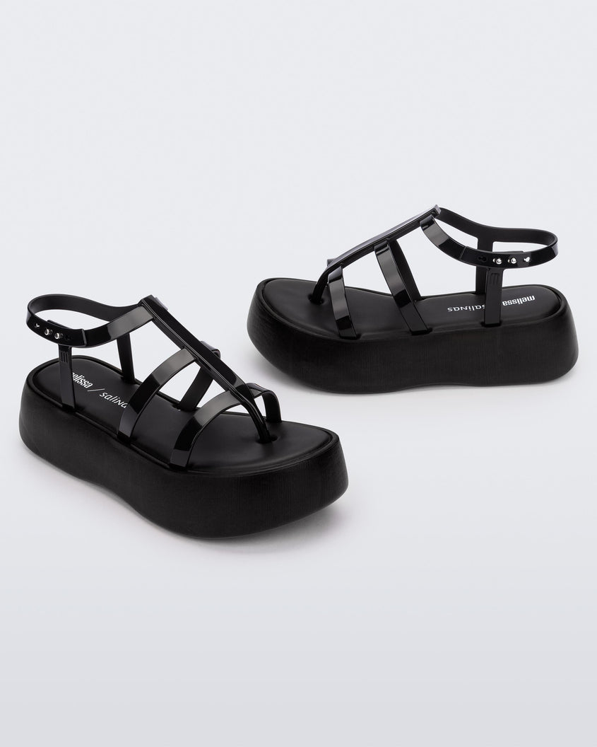 An angled front and side view of a pair of black Melissa Caribe High Platform sandals with two black front straps joined by a vertical strap intersecting the ankle strap and a black sole.