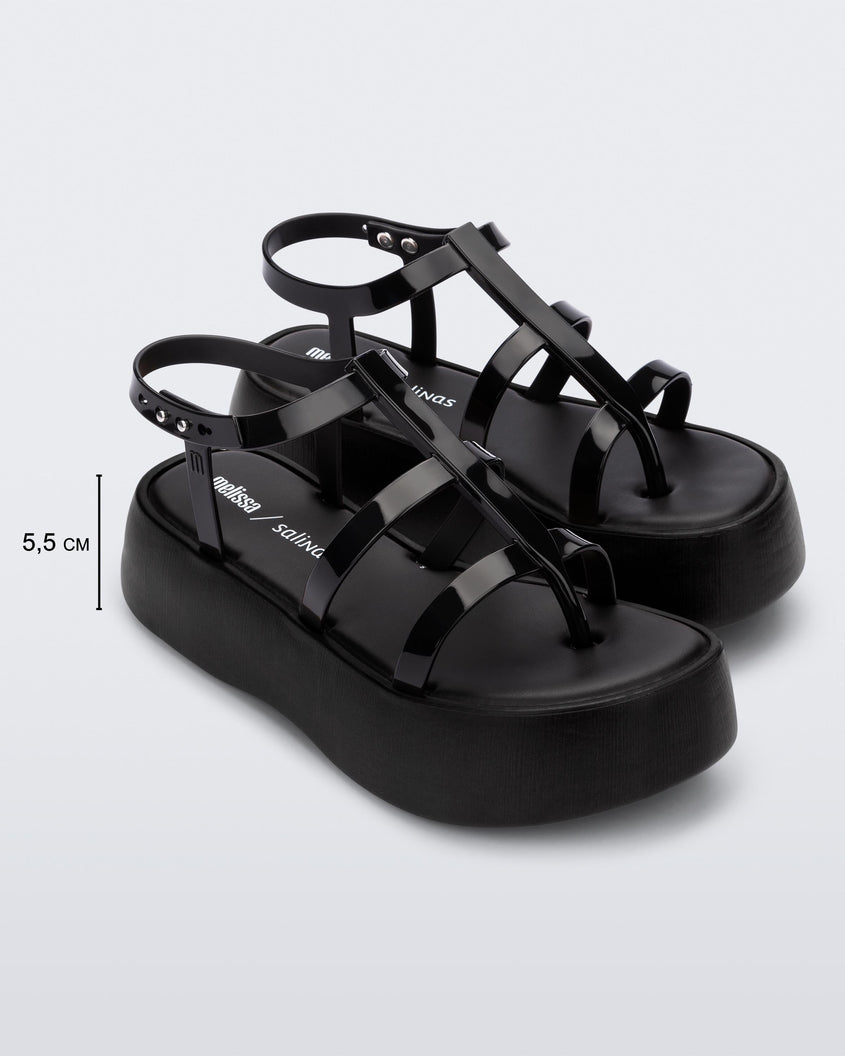 An angled side view of a pair of black Melissa Caribe High Platform sandals with two black front straps joined by a vertical strap intersecting the ankle strap and a black sole, showing the dimensions of the platform which read 5.5 centermeters high.