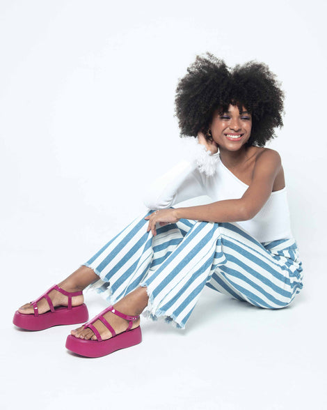 A model sitting on the ground, posing for a picture in a white top, blue and white striped bottoms and a pair of pink Melissa Caribe High Platform sandals with two pink front straps joined by a vertical strap intersecting the ankle strap and a black sole.