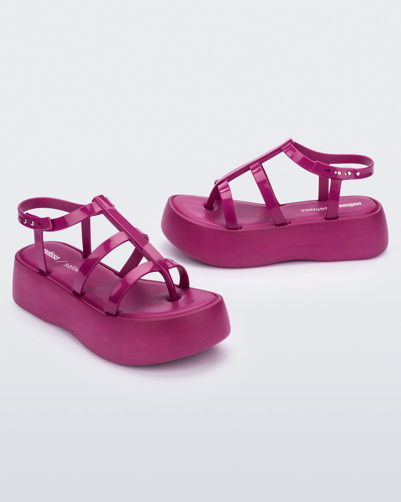 An angled front and side view of a pair of pink Melissa Caribe High Platform sandals with two pink front straps joined by a vertical strap intersecting the ankle strap and a black sole.